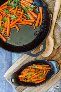 two cast iron pans of completed maple glazed carrots, one resting on a cutting board with a wooden spoon