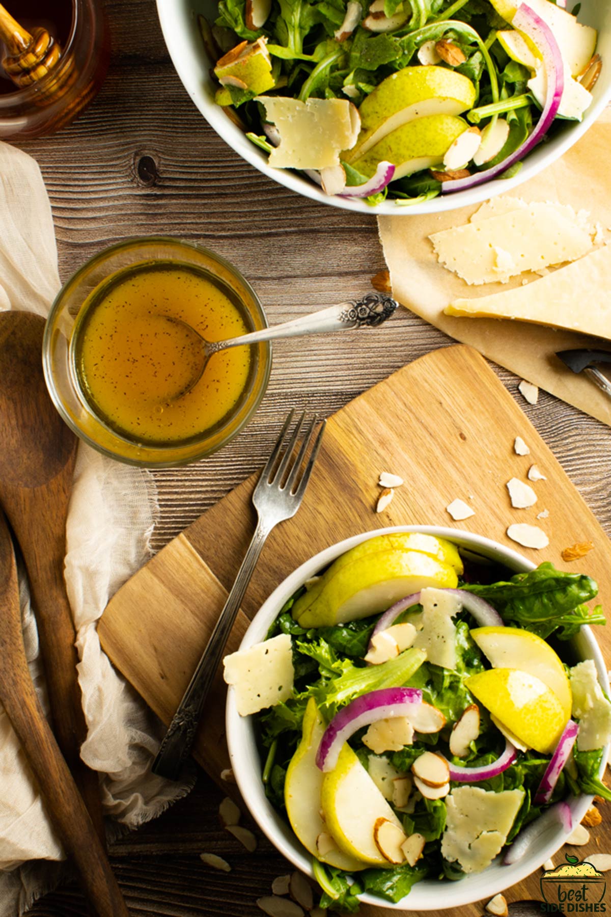 a bowl of salad on a cutting board next to a dish of vinaigrette, cheese and utensils