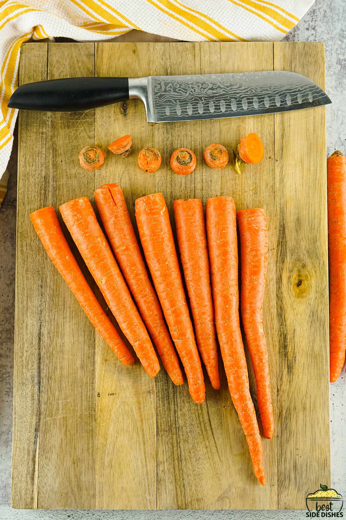 carrots lined up on a wooden cutting board with the tops cut off, next to a knife