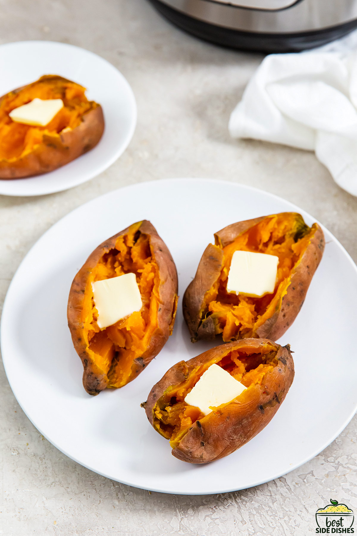 three sweet potatoes on a plate with cut open skin and a pat of butter on each potato, next to another plate with another sweet potato.