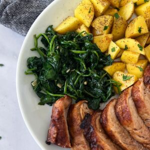 sautéed spinach on a white plate with seasoned potatoes and sliced pork