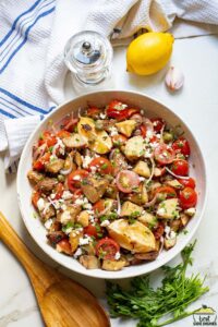 white bowl of greek potato salad next to a wooden spoon, herbs, lemon and shallots