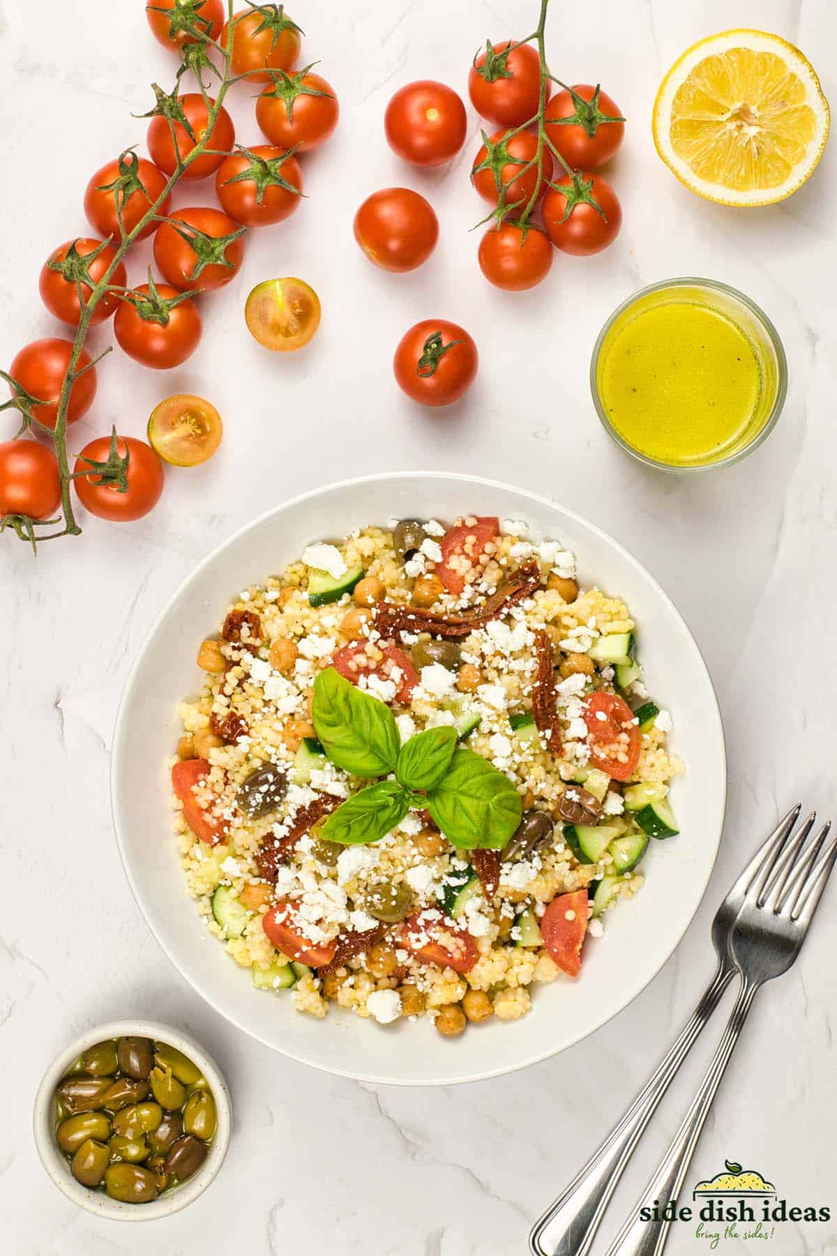 a bowl of mediterranean couscous salad next to fresh tomatoes, olives, olive oil and a lemon