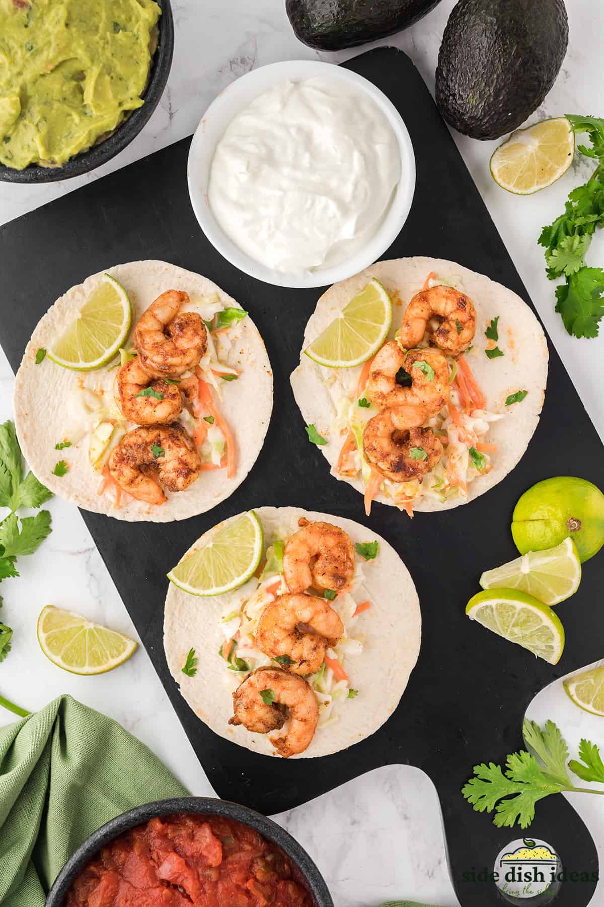 three shrimp tacos with coleslaw on a platter with sour cream in a bowl and lime slices