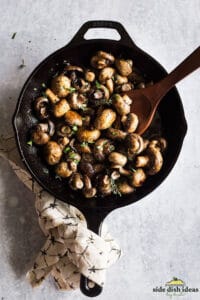 stirring white button mushrooms with a wooden spoon in a skillet