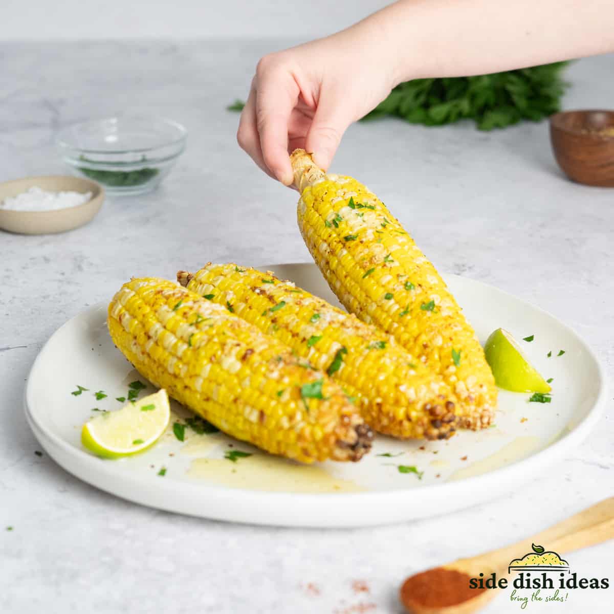 Three pieces of air fried corn on the cob on a white platter with limes and herbs